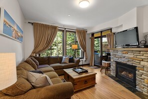 Ein Wohnzimmer mit Ecksofa, Kamin, Fernseher und einem Fenster mit Vorhängen.