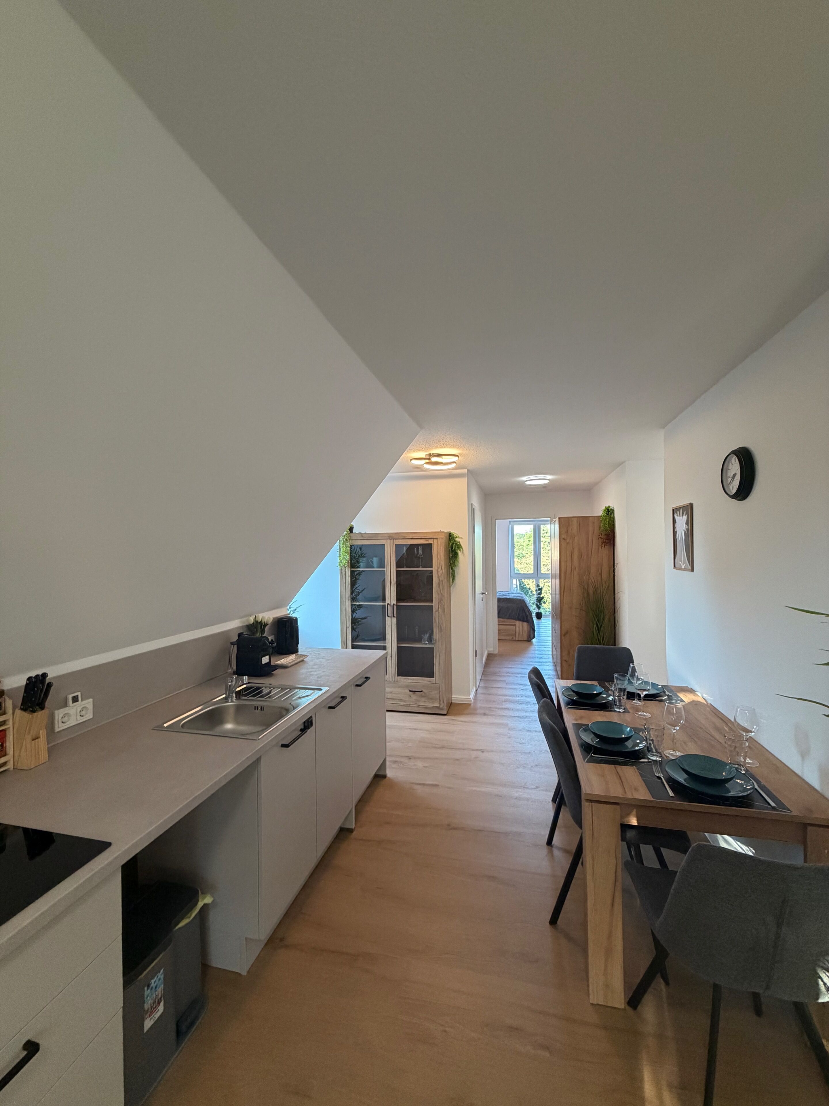 Eine moderne Küche mit einer zentralen Kochinsel, ein Essbereich mit Holztisch und -stühlen sowie Blick in das Wohnzimmer durch eine offene Tür.