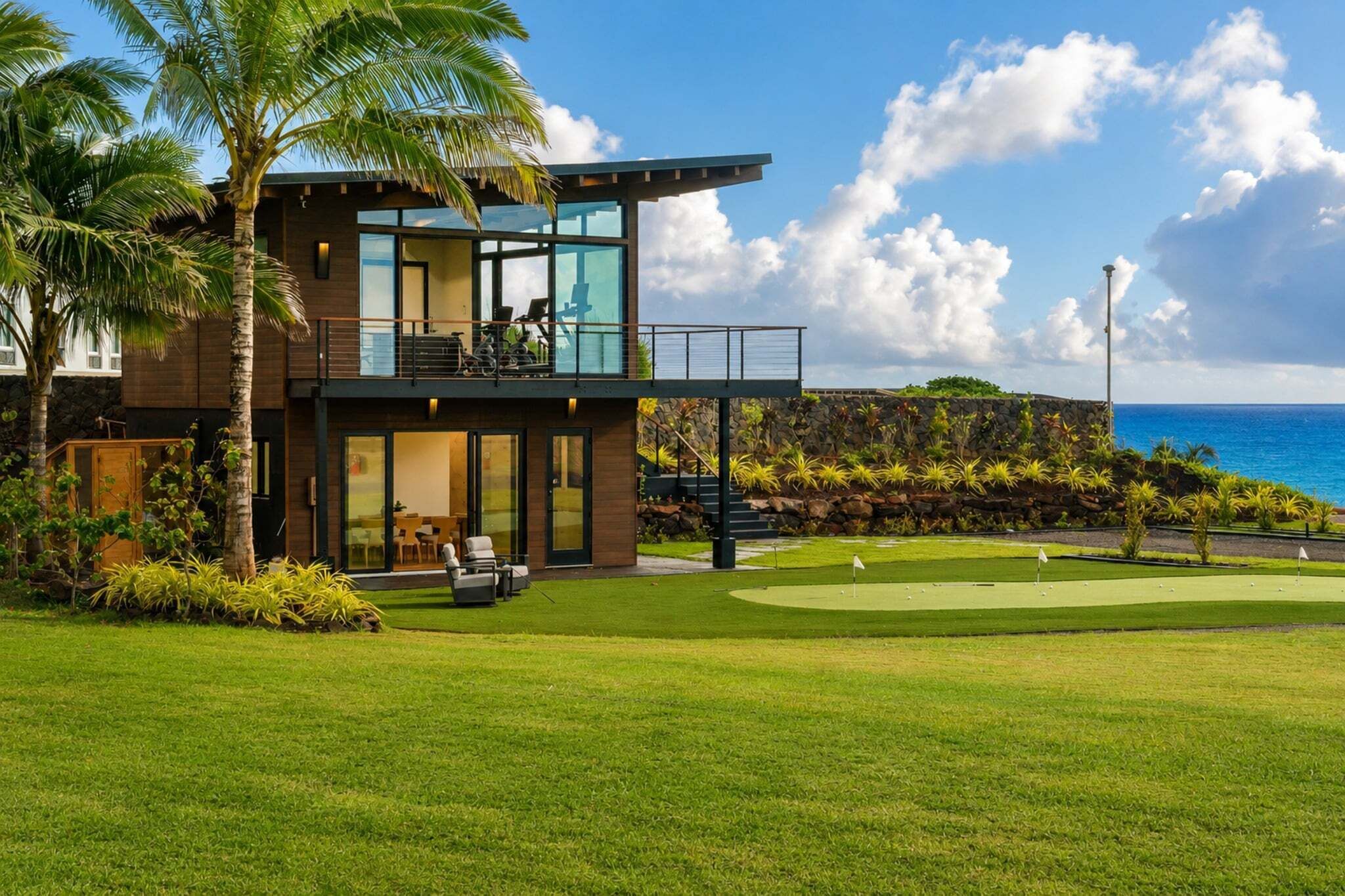 A modern two-story house with a balcony, a patio with outdoor furniture, and a view of the ocean.