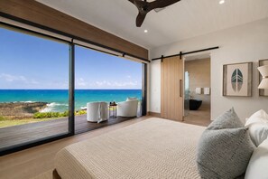 Ein Schlafzimmer mit großem Fenster und Blick auf einen Strand, einem Bett mit Kissen, einem kleinen Tisch und einem Stuhl.