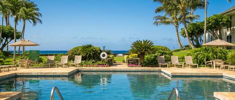 A pool with clear blue water, surrounded by lounge chairs and palm trees, with a view of the ocean in the background.