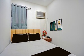 Un dormitorio con cama, una mesita de noche con teléfono y un florero, aire acondicionado instalado en la pared, y una ventana con cortina.