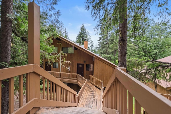 A wooden cabin with a wooden staircase leading to a deck, surrounded by tall trees. - Limber Pine Townhome and 2bdr Stay (Whitefish)