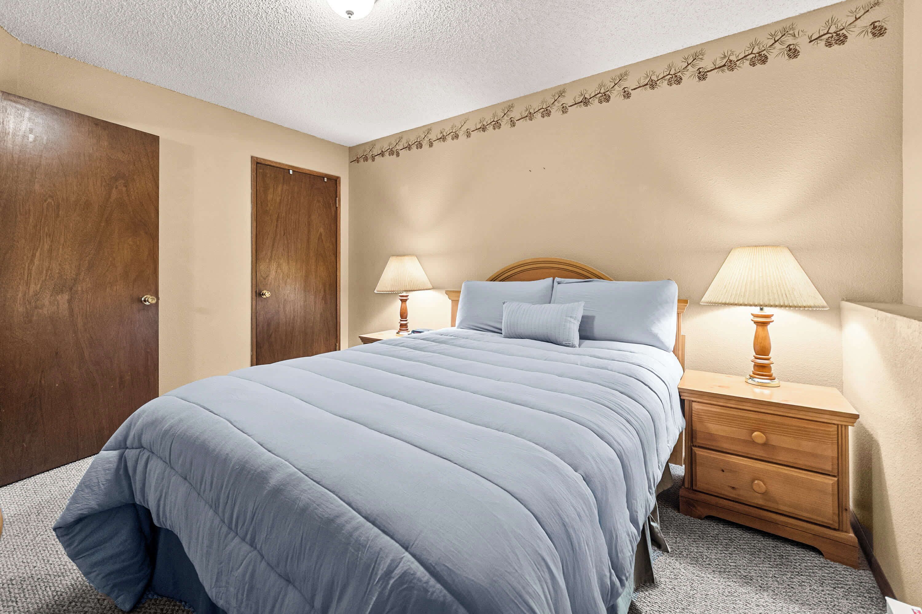 A bedroom with a large bed, two bedside tables with lamps, a wooden door, and a wall with a pine branch decoration.