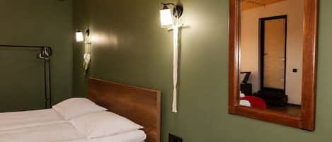 A room with a bed, a wooden headboard, a wall-mounted light fixture, and a mirror reflecting a window with blinds.