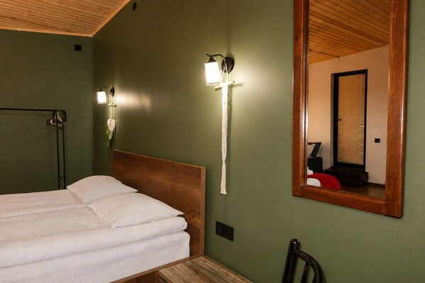 A room with a bed, a wooden headboard, a wall-mounted light fixture, and a mirror reflecting a window with blinds.