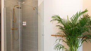 A glass-enclosed shower with a gold showerhead and handheld showerhead, a white towel on the floor, and a potted plant to the right. - Deluxe Quadruple Apartment (Alicante)