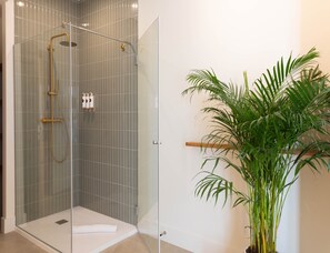 A glass-enclosed shower with a gold showerhead and handheld showerhead, a white towel on the floor, and a potted plant to the right. - Deluxe Quadruple Apartment (Alicante)