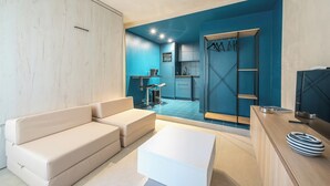 A modern living room with a sofa, a coffee table, and a TV. There is a kitchen area visible in the background with blue cabinets and a sink. - Brand New Modern Studio in Faliro - 
Private garden (Paleo Faliro)