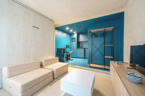 Ein modernes Wohnzimmer mit einer Couch, einem Couchtisch und einem Fernseher. Im Hintergrund ist eine Küche mit blauen Schränken und einer Spüle erkennbar.