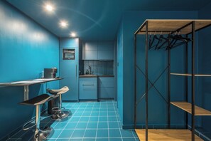 A compact kitchen with blue walls, a coffee machine, a sink, and a small dining area with a chair. - Brand New Modern Studio in Faliro - 
Private garden (Paleo Faliro)