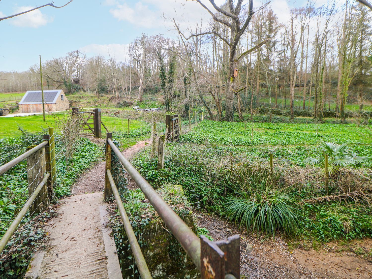 Eine rustikale Holzbrücke über einen Feldweg, der zu einem grünen Feld mit einer Scheune im Hintergrund führt.