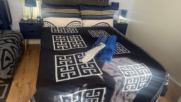 A neatly made bed with a geometric patterned bedspread, a folded towel, and a bedside lamp on a nightstand. - Cozy London Apartment With View (London)