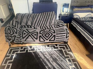 Ein Zimmer mit einem Bett, einem Sessel, einem Nachttisch und einem Teppich mit geometrischem Muster.