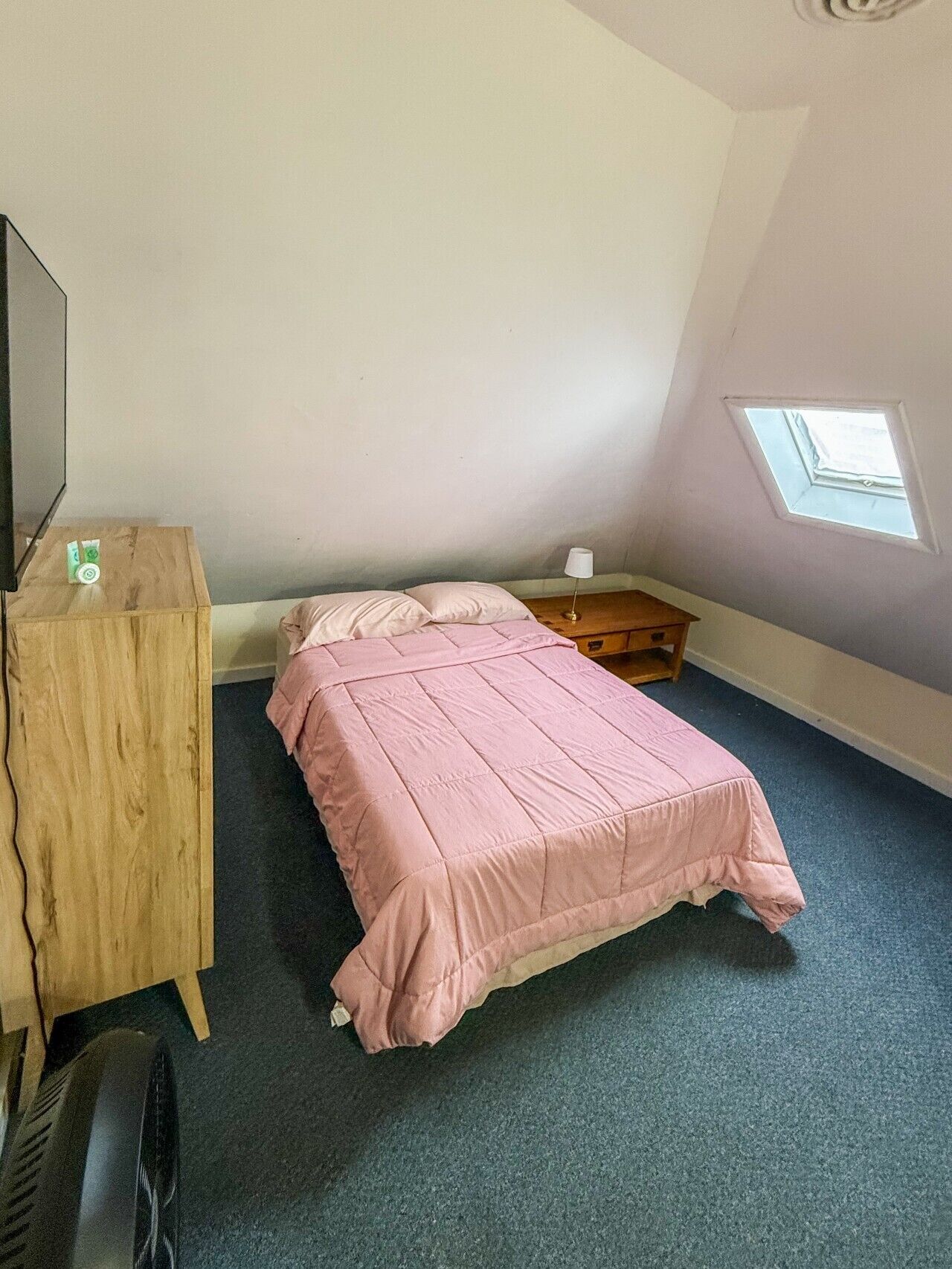 A bedroom with a bed, a dresser, a desk, a lamp, and a skylight.