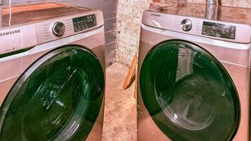 Two Samsung washing machines in a laundry room with a brick wall and wooden shelving.
