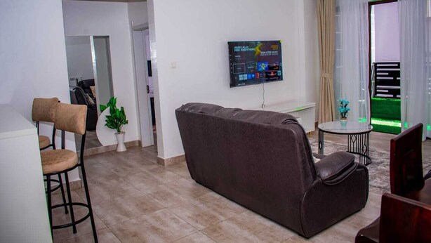 Una sala de estar con sofá, televisión montada en la pared, una mesita redonda y una planta en maceta.