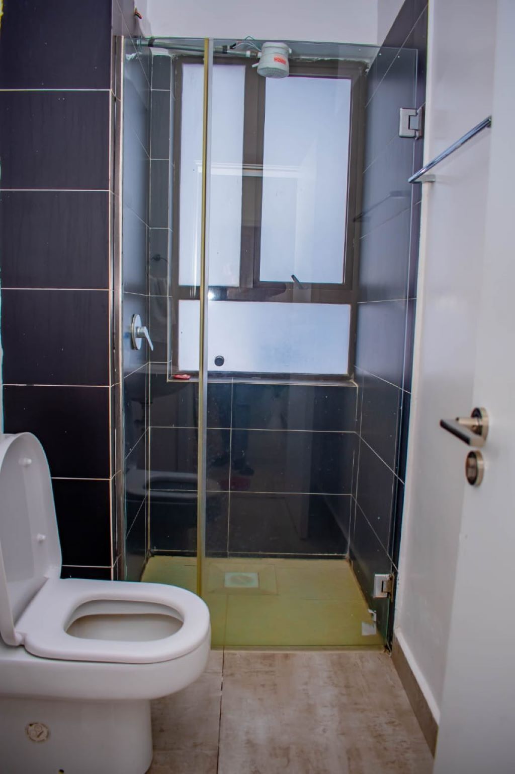 Um espaço de banho pequeno e fechado, com divisória de vidro, vaso sanitário e chão revestido de azulejos.