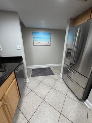 A kitchen with a stainless steel refrigerator, a granite countertop, and a painting of a beach scene on the wall. - Safe & Sound (Biloxi)