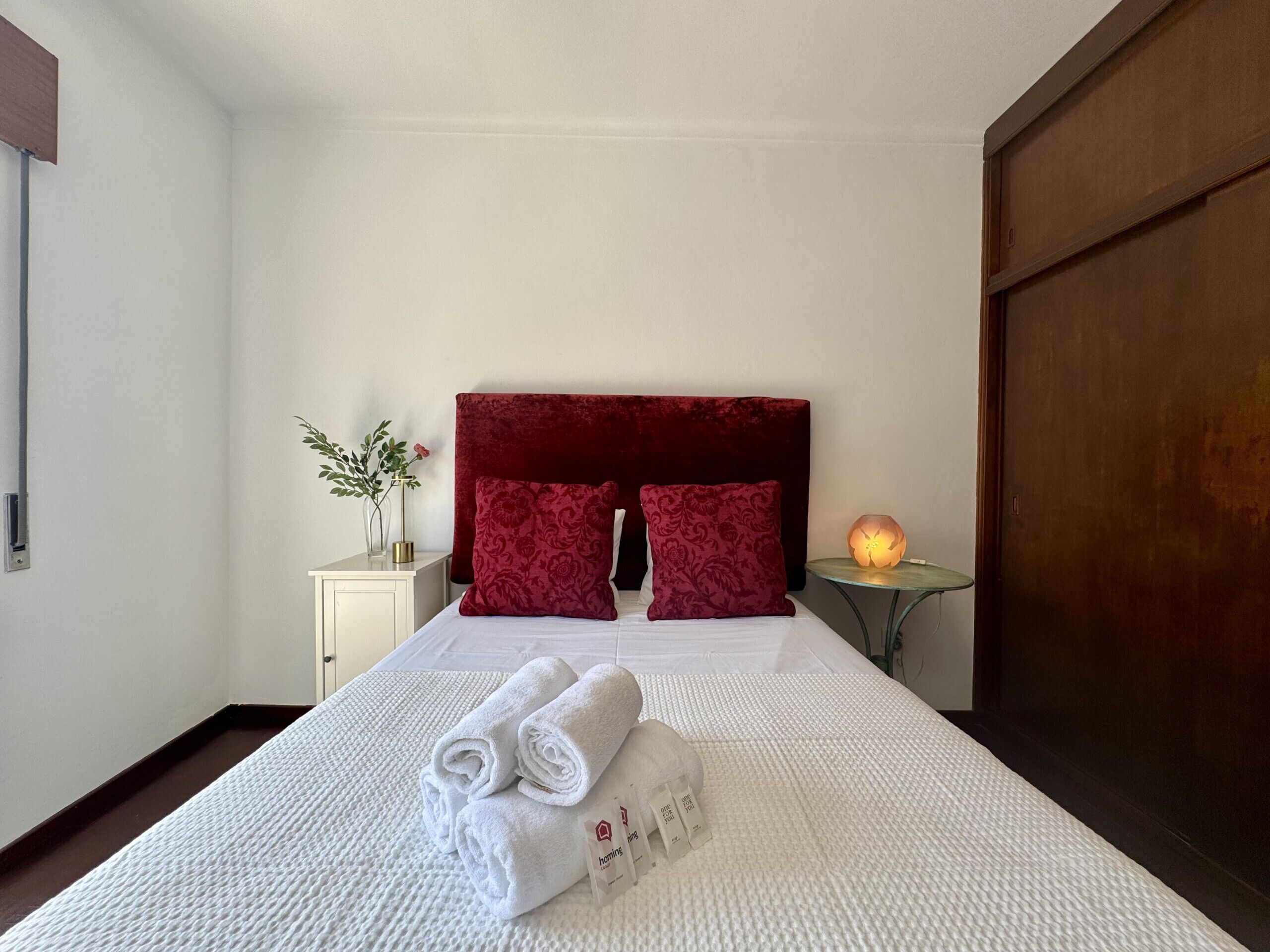 Une chambre d’hôtel avec un lit, des oreillers, une table de chevet avec un vase et une petite table de nuit avec une lampe.