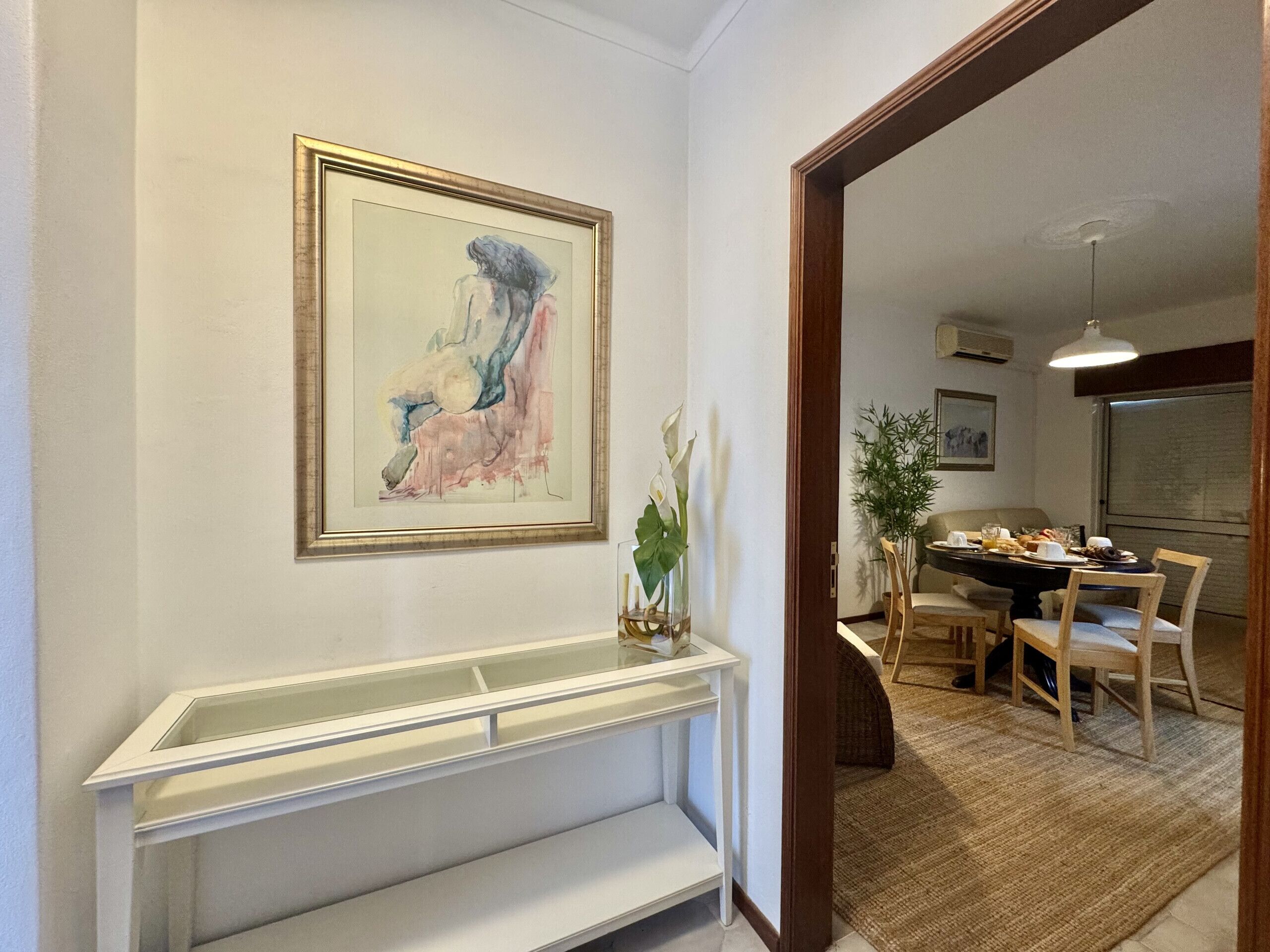 A hallway with a framed abstract painting, a white console table, and a dining area with chairs and a table set for a meal.