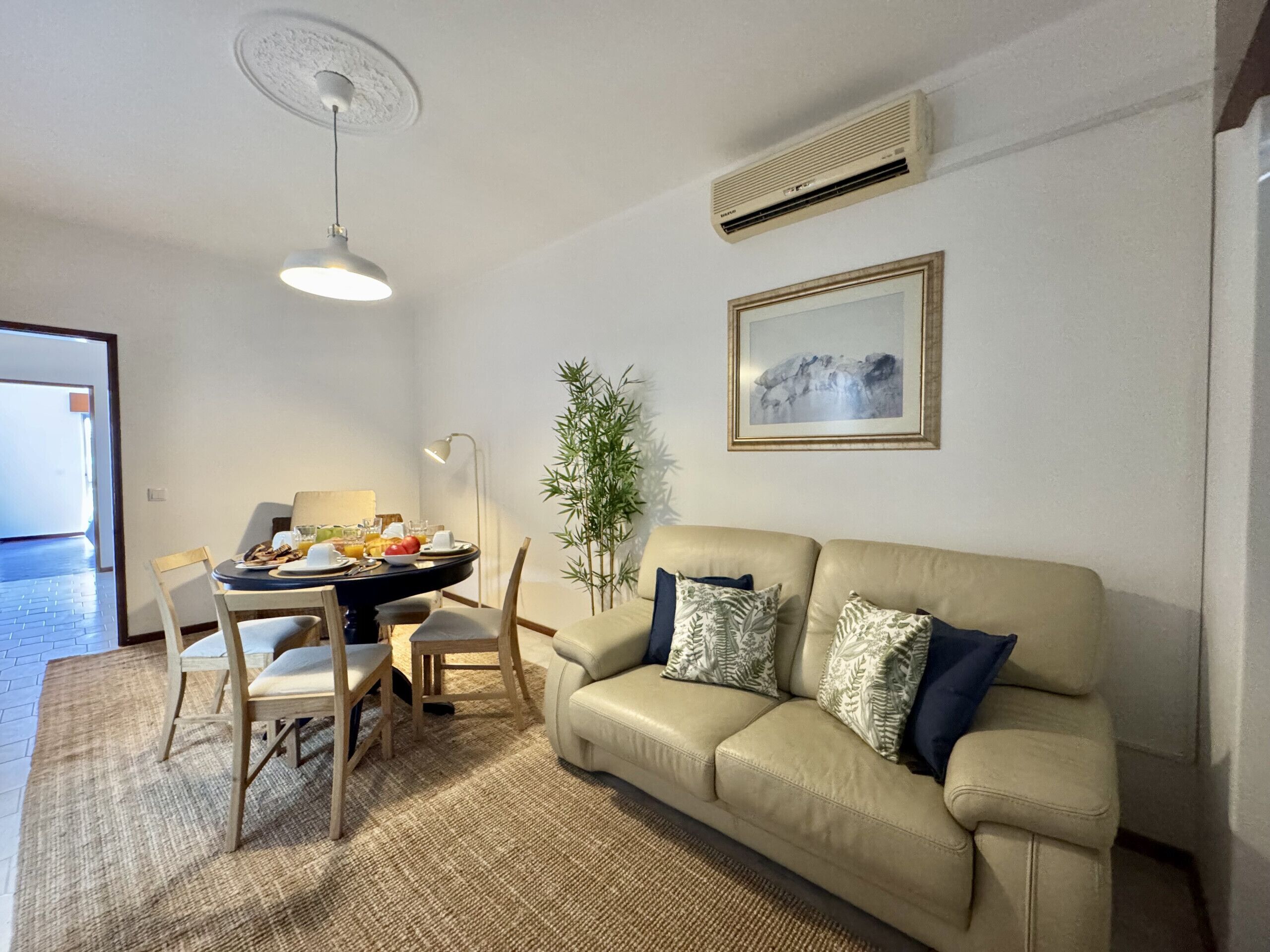 A living room with a beige sofa, a dining table with chairs, a floor lamp, and a potted plant.