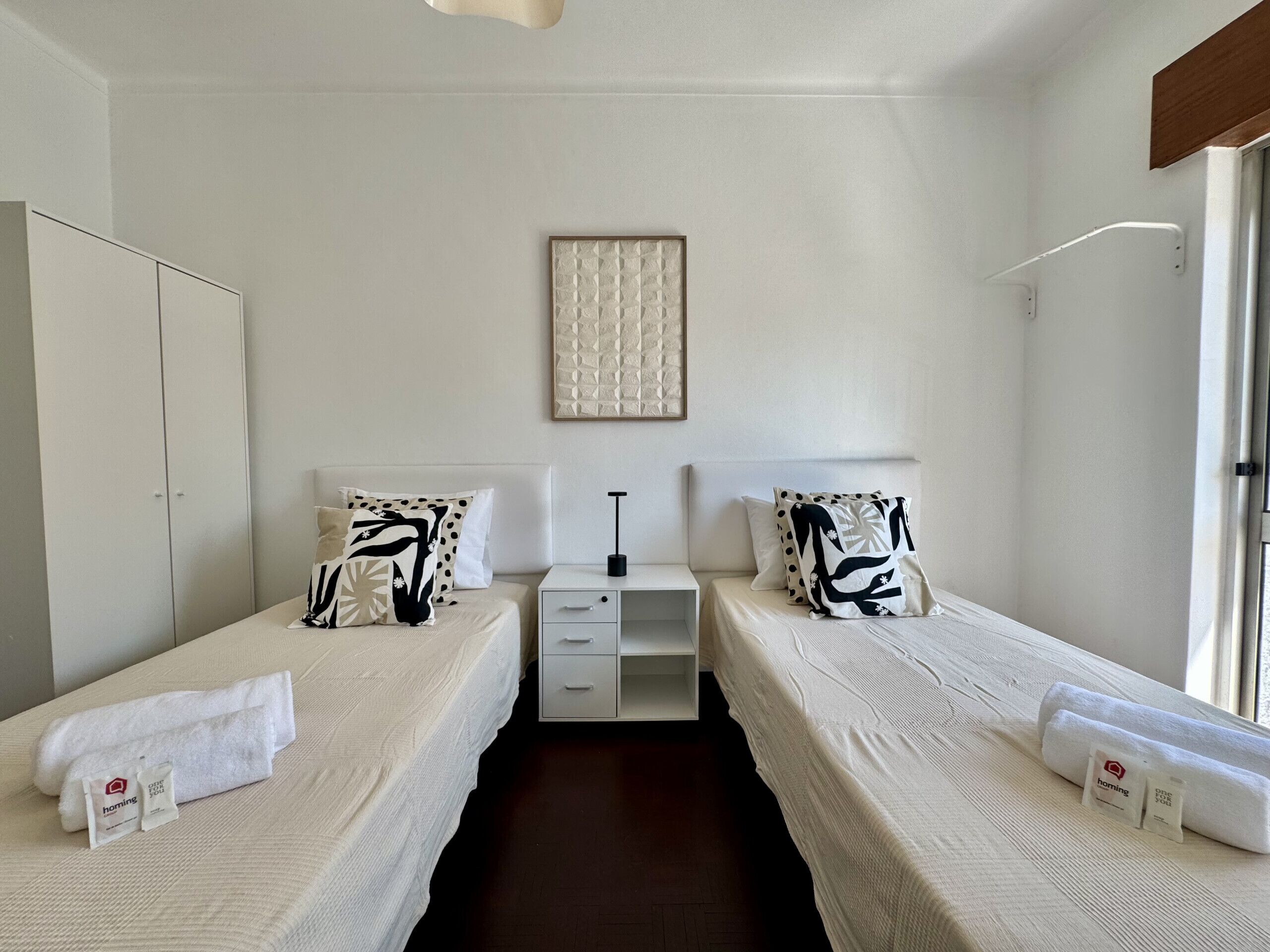 Habitación de hotel con dos camas individuales, una mesita de noche con lámpara, un armario y un cuadro en la pared.