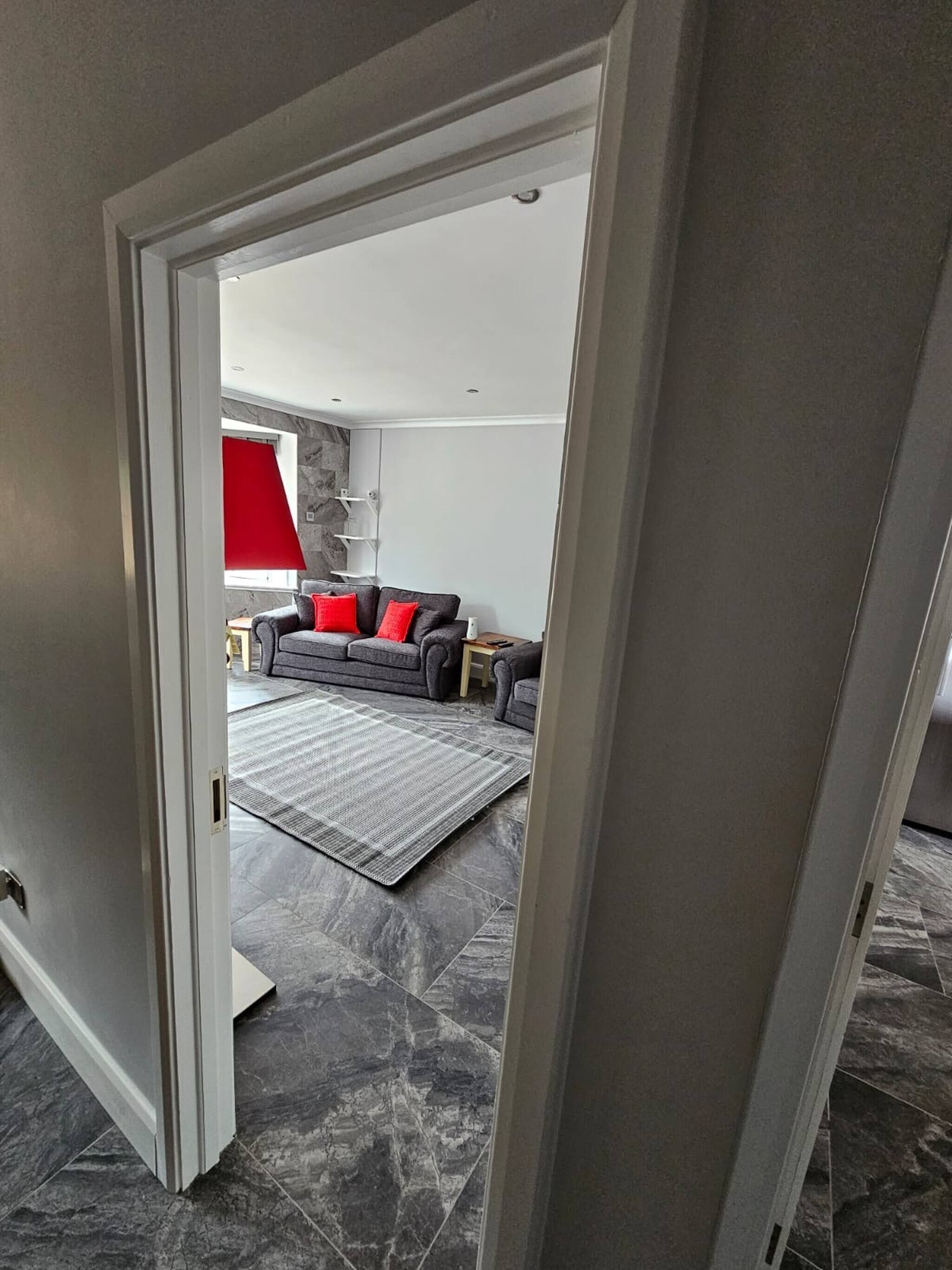 A modern living room with a grey sofa, red cushions, a patterned rug, and a stone-tiled floor.
