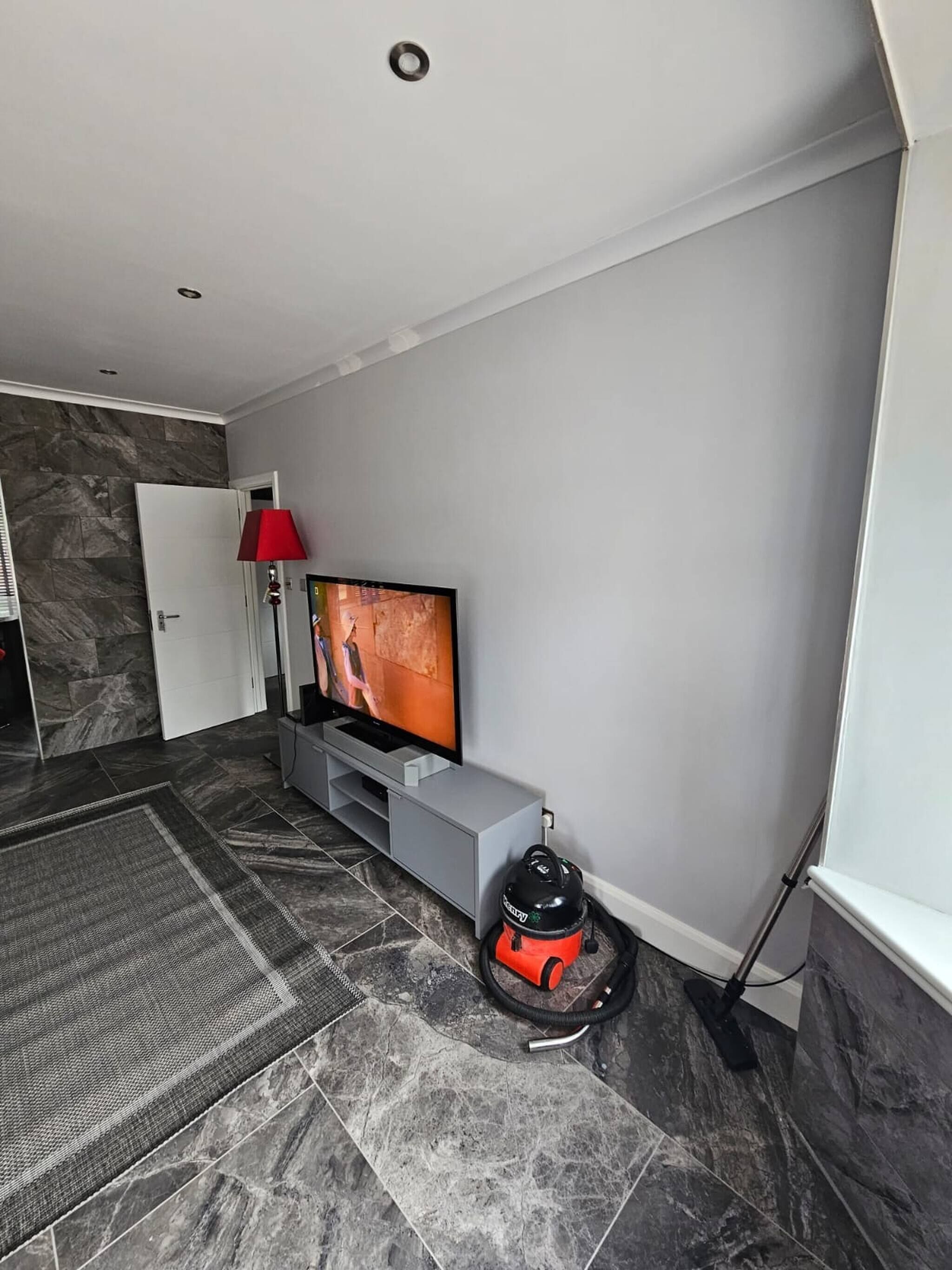 A modern living room with a flat-screen TV, a red floor lamp, and a vacuum cleaner on the floor.
