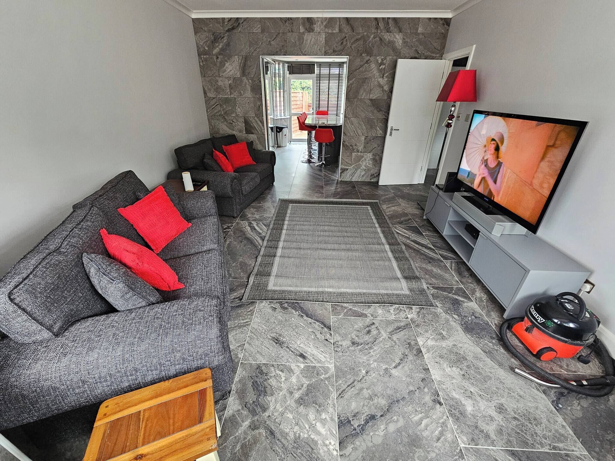 Una sala de estar moderna con un sofá gris, cojines rojos, un televisor y una aspiradora.