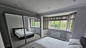 A bedroom with a bed, a large mirror, and a window with blinds. - Serene 5-bedroom House in Staines-upon-thames With Garden (Staines)