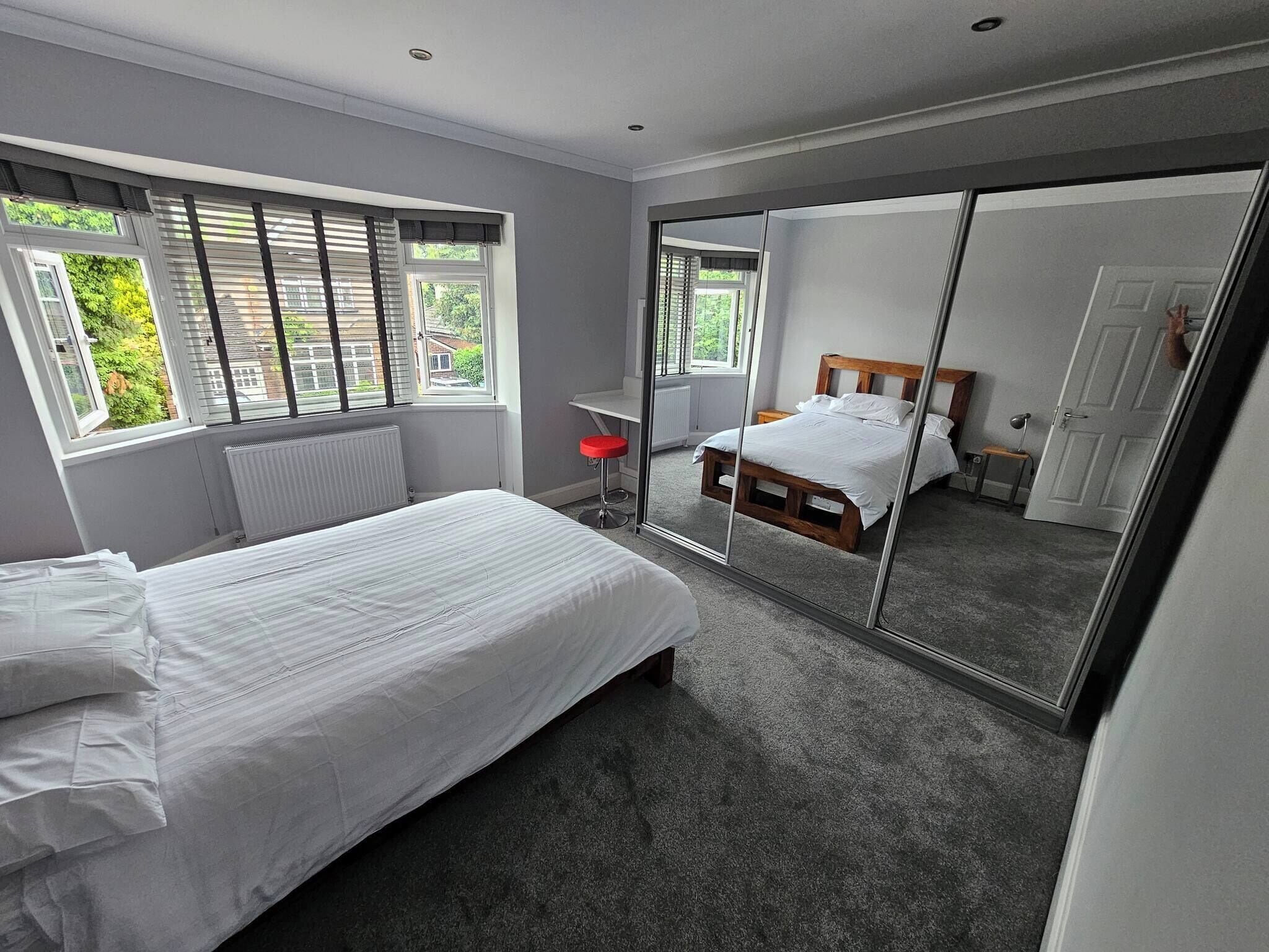 A bedroom with a bed, a red stool, a large mirror, and a view of a garden through the window.