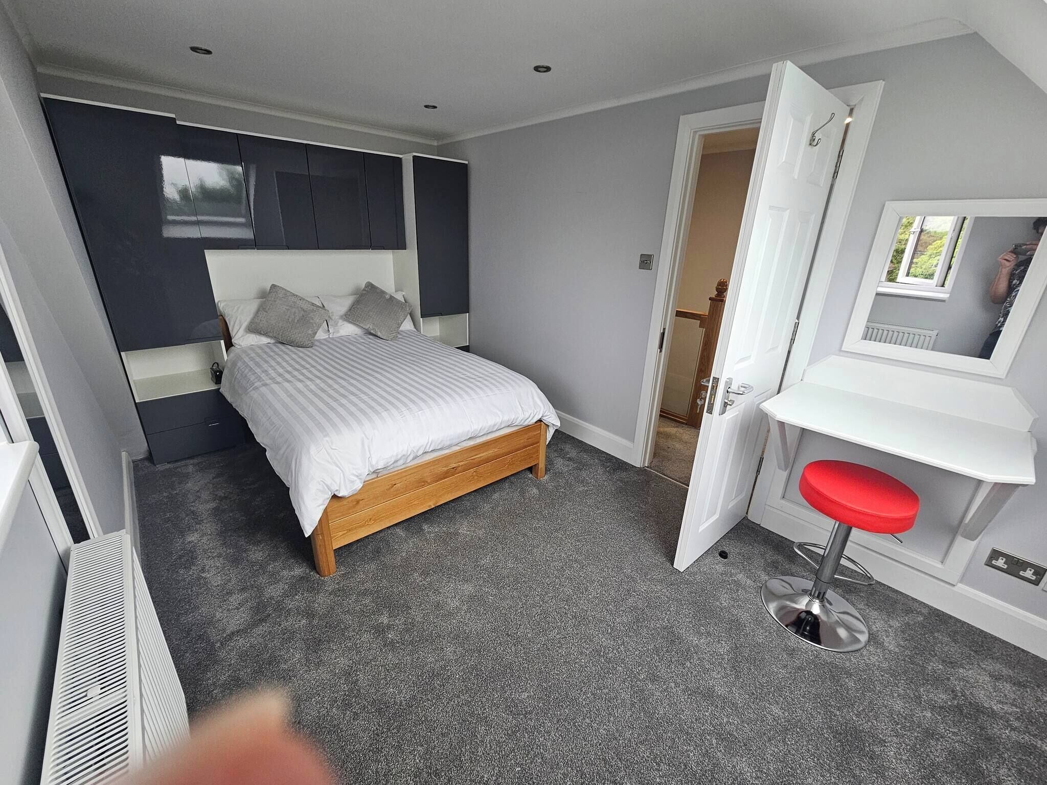 A bedroom with a bed, a desk with a red chair, and a door leading to another room.