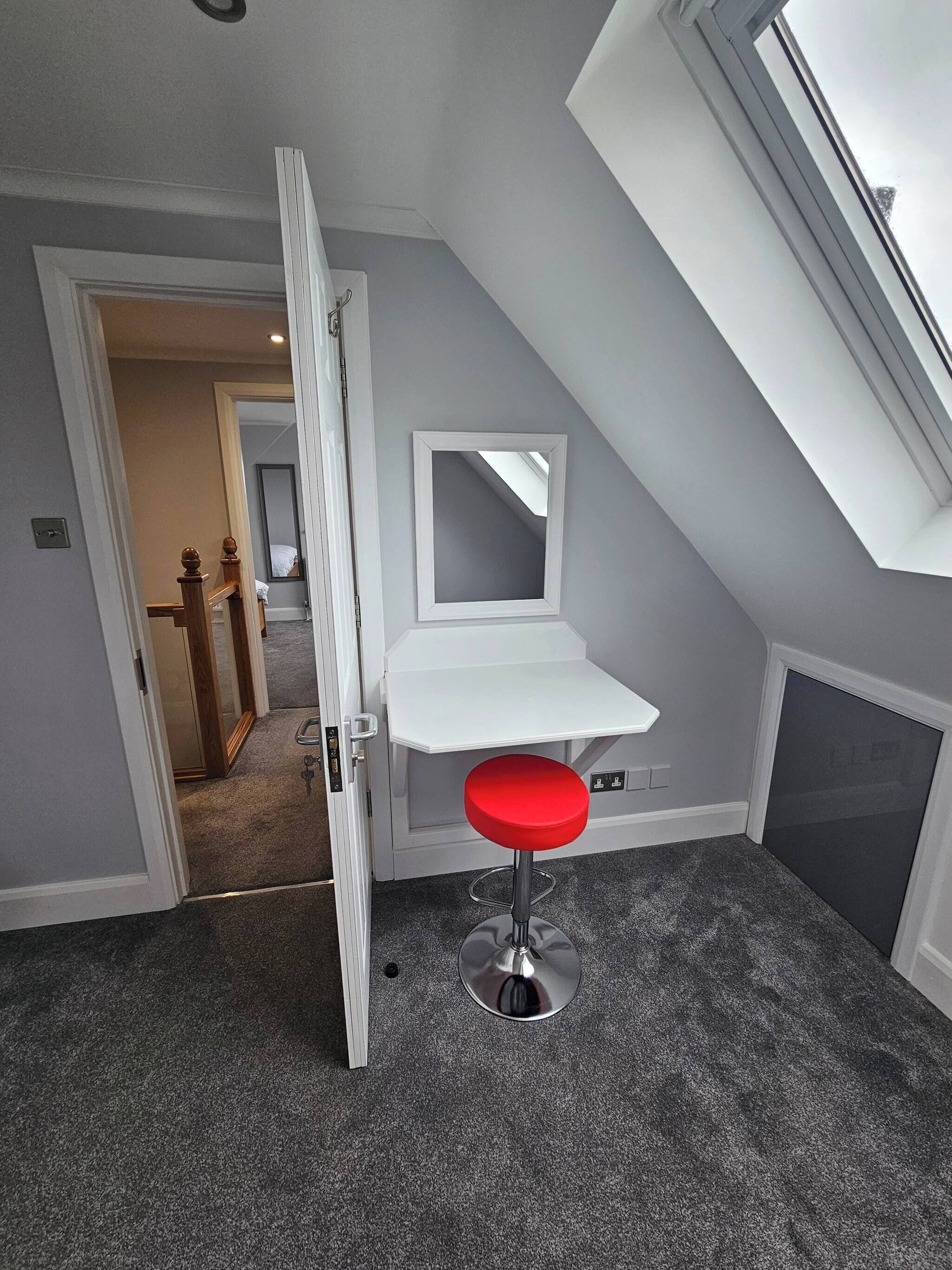 A room with a sloped ceiling, a white desk with a mirror, a red stool, and a door leading to another room.