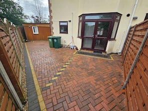 Un patio empedrado con diseño de ladrillo, cerca de madera y un edificio con puerta de marco oscuro.