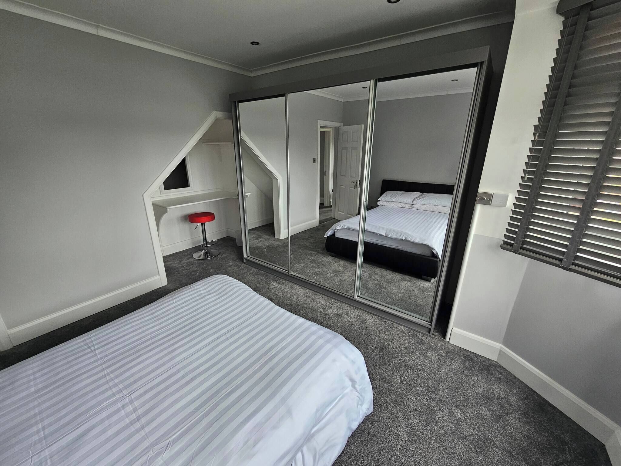 A bedroom with a bed, a red stool, a large mirror, and a staircase leading to another room.
