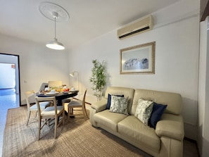 A living room with a beige sofa, a dining table with chairs, a floor lamp, and a potted plant. - FARO CENTRAL 1 by HOMING (Faro)