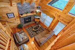 A cozy cabin interior with a stone fireplace, wooden walls, a ceiling fan, and a large window. - The Cats' Lair: Spectacular Views, Hot Tub, Wifi, Game Room, Resort Pool (Pigeon Forge)