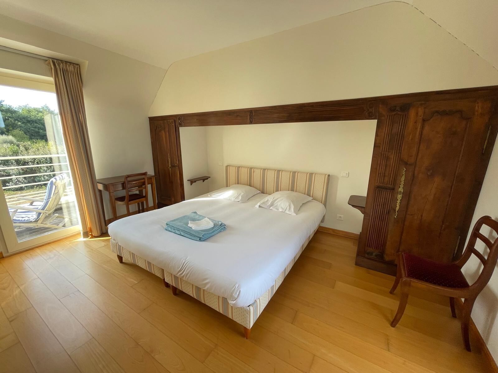 A bedroom with a large bed, wooden headboard, a small table, a chair, and a view through a glass door.