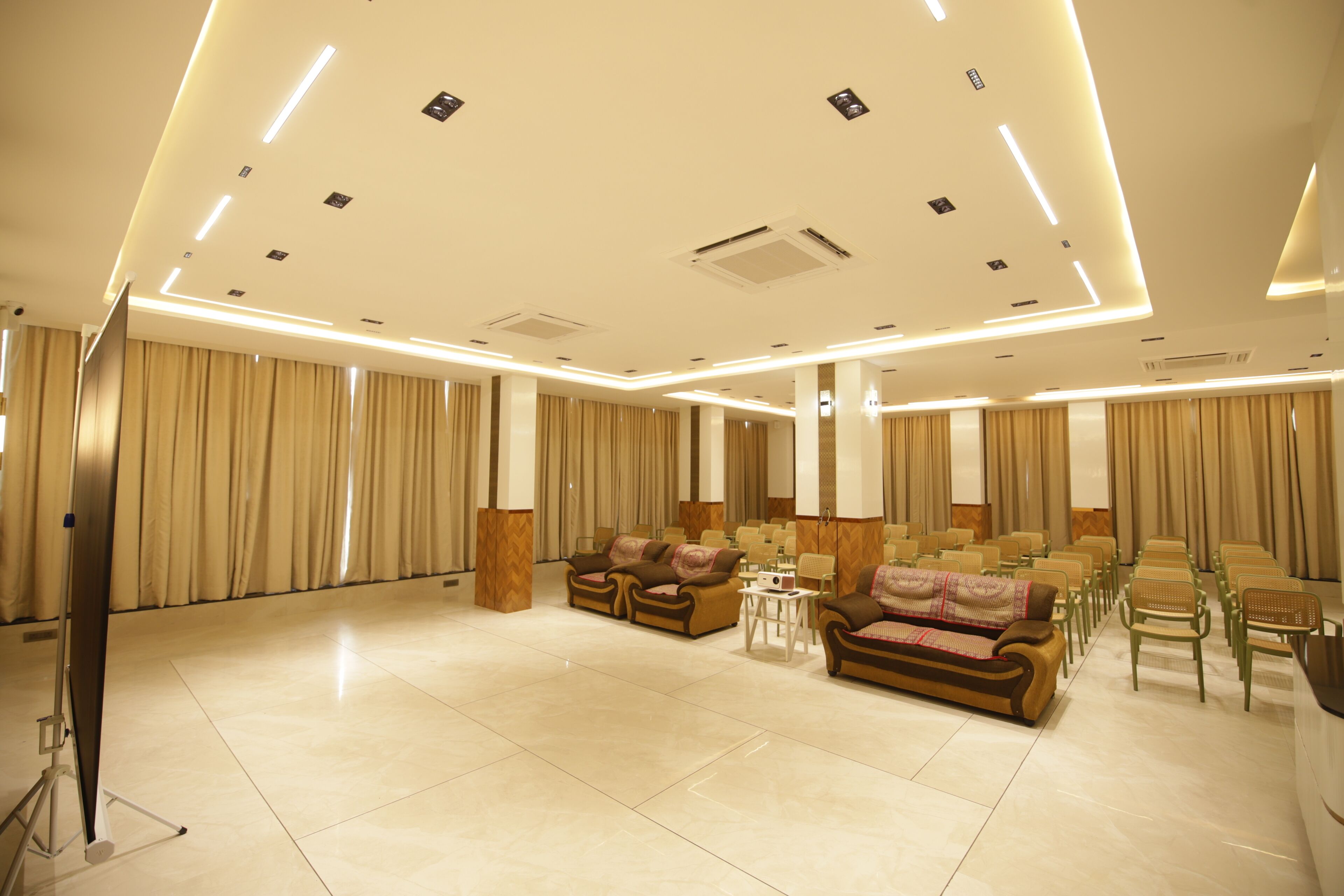 A spacious hotel lobby with a high ceiling, recessed lighting, a seating area with sofas and chairs, and a large window with curtains.