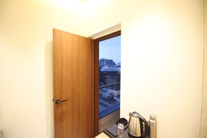 A room with a wooden door, a window with a view of buildings, a kettle, and a water bottle. - Fort View Palace (Lachhmangarh)
