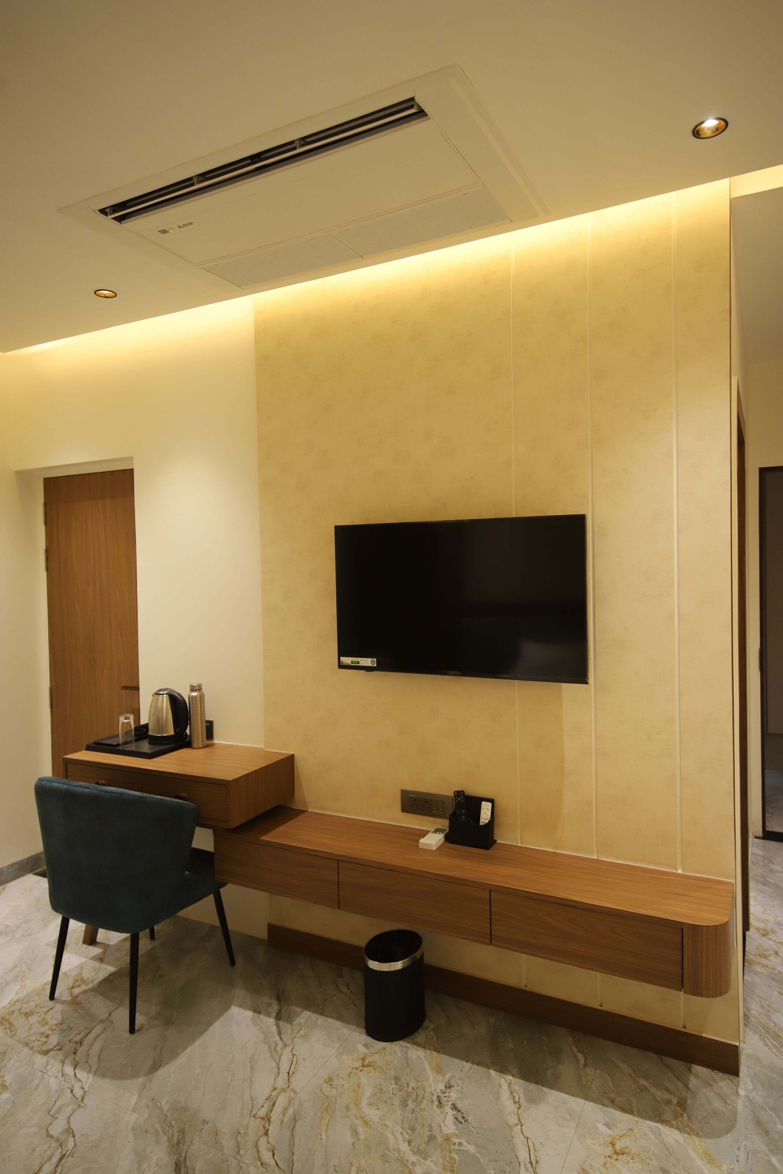 A modern hotel room with a flat-screen TV mounted on a wall, a wooden desk with a chair, a coffee maker, and a remote control on the desk.