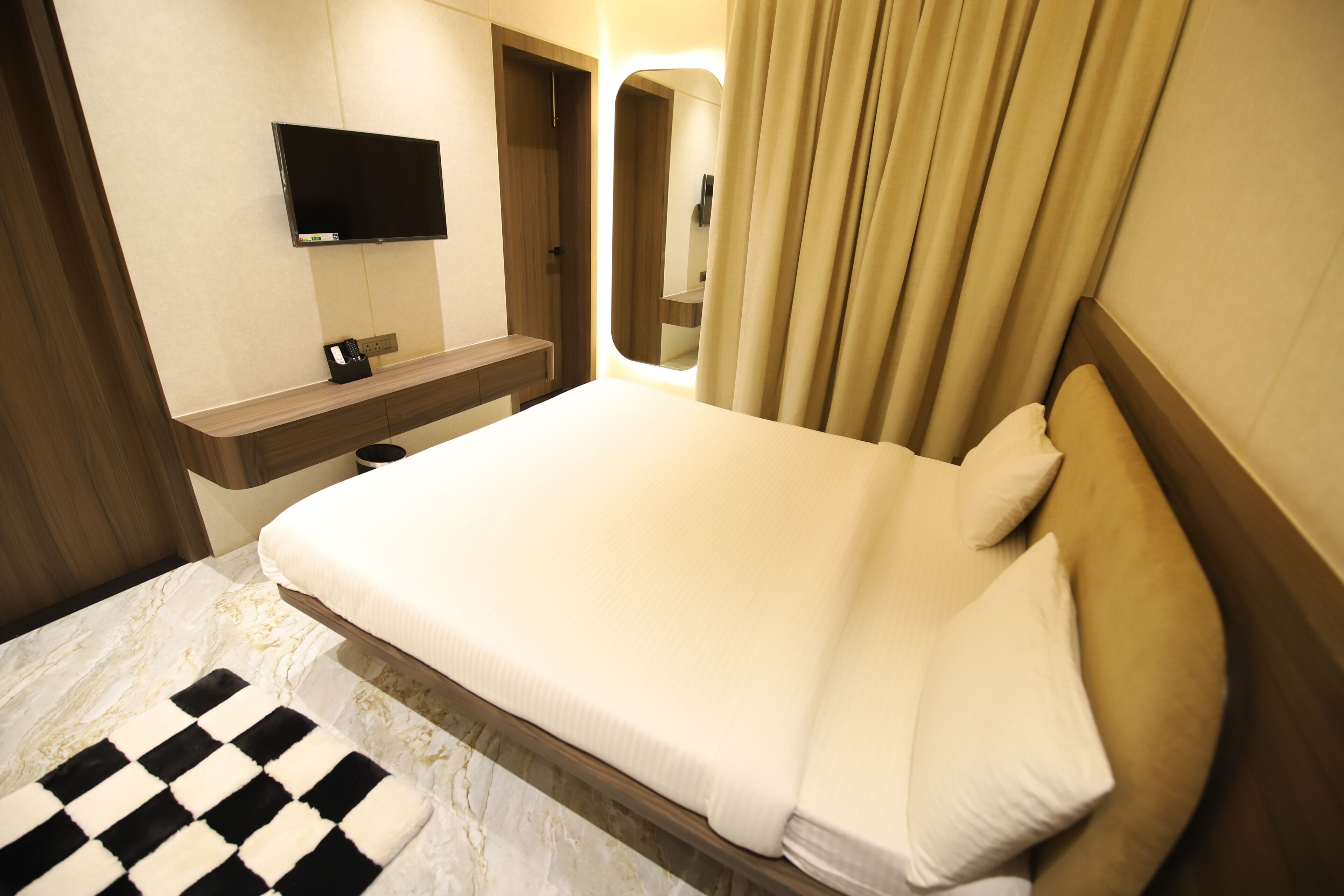Habitación de hotel con cama, televisor de pantalla plana montado en la pared, cabecera de madera y baño con espejo y cortinas.