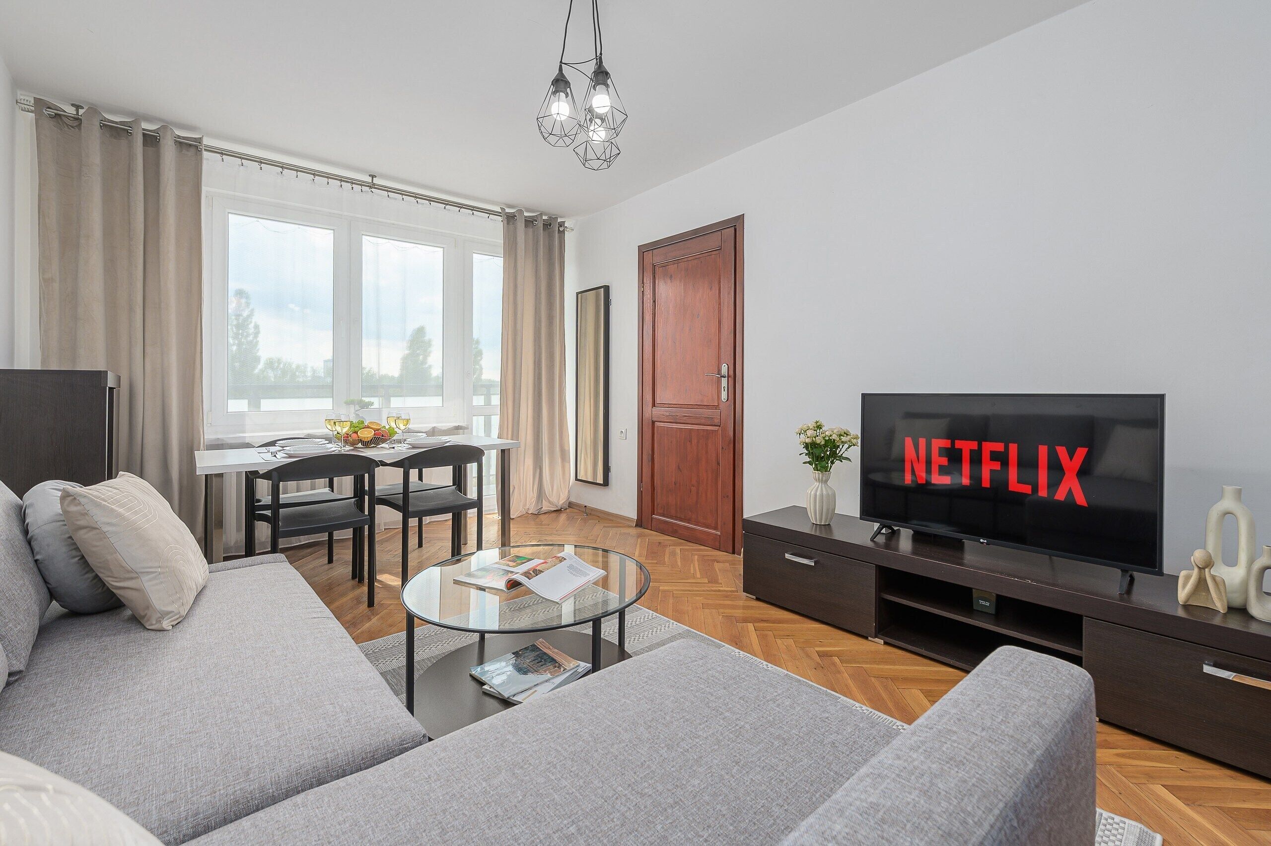 A modern living room with a sofa, coffee table, dining table, and a television displaying the Netflix logo.