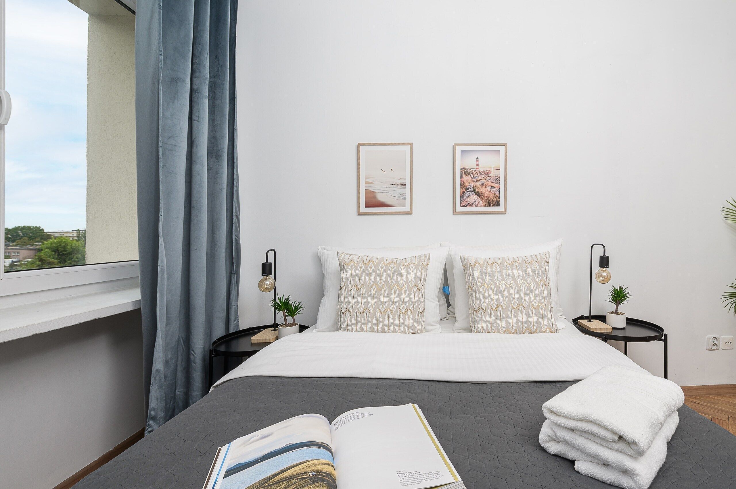 A hotel room with a bed, two bedside tables, a window with curtains, and two framed pictures on the wall.