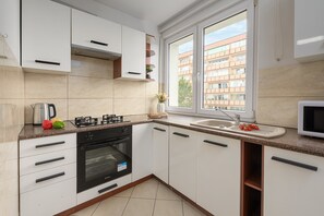 A modern kitchen with white cabinets, a gas stove, an oven, a microwave, and a window with a view of buildings. - Lumina 3room Apart with Balcony near ZOO (Warszawa)