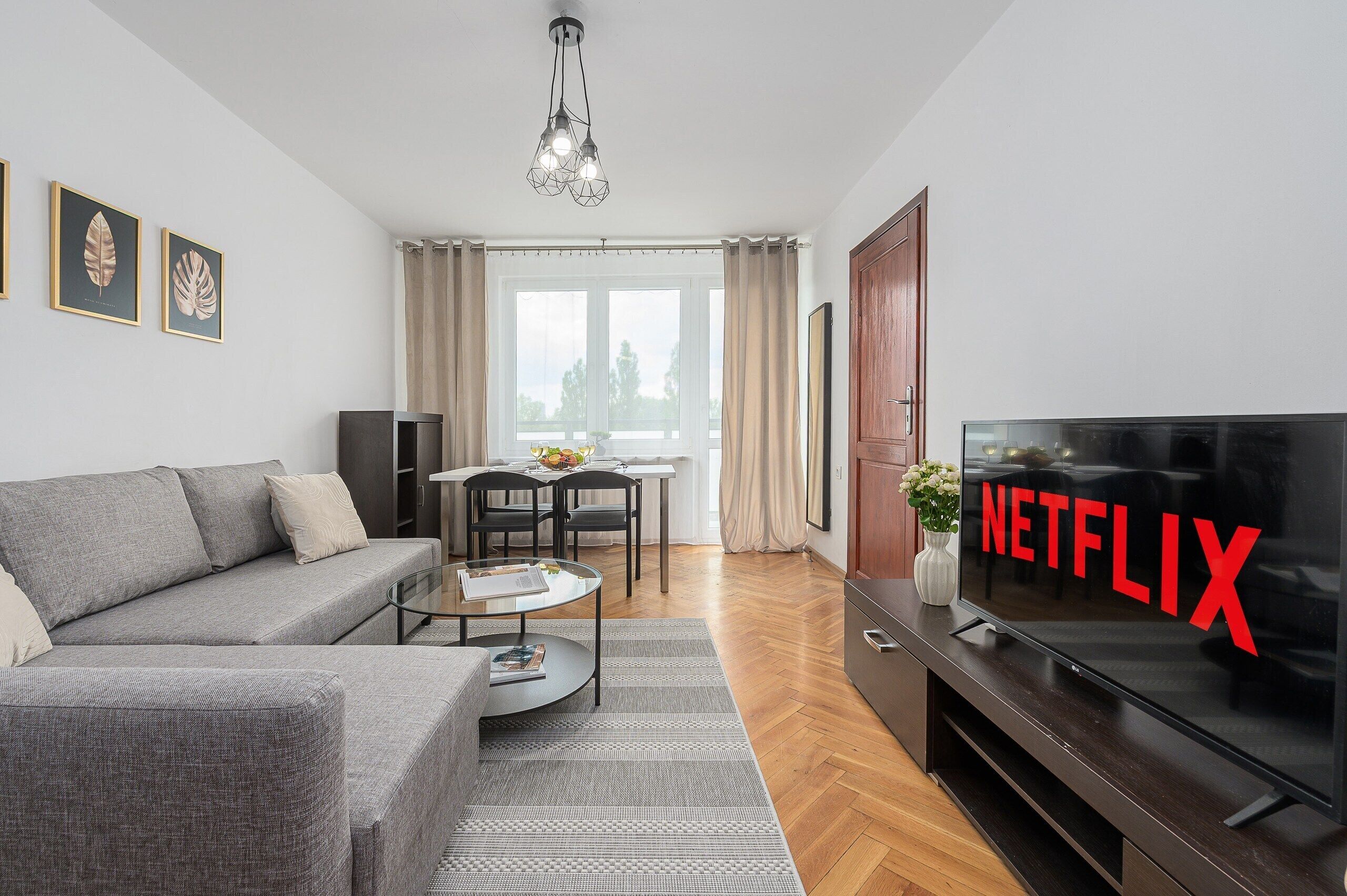 Un salon moderne avec un canapé gris, une table basse, un téléviseur orné du logo Netflix et un coin repas comprenant une table et des chaises.