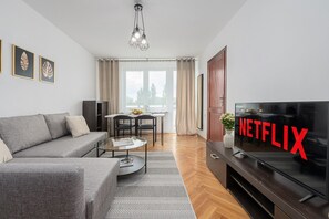 A modern living room with a grey sofa, a coffee table, a TV with a Netflix logo, and a dining area with a table and chairs. - Lumina 3room Apart with Balcony near ZOO (Warszawa)