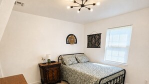 Ein Schlafzimmer mit Bett, Kommode, Nachttisch, Wandlampe, Deckenleuchte und einem Fenster mit Jalousien.