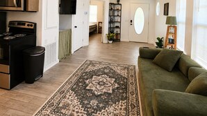 Ein großzügiger Wohnbereich mit hoher Decke, eine moderne Küche mit integrierten Geräten, ein großes Fenster und ein gemütliches, grünes Sofa.